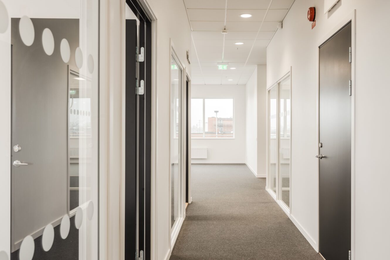 En modern kontorskorridor med grå matta, vita väggar, flera stängda dörrar och stora fönster i slutet som släpper in naturligt ljus.