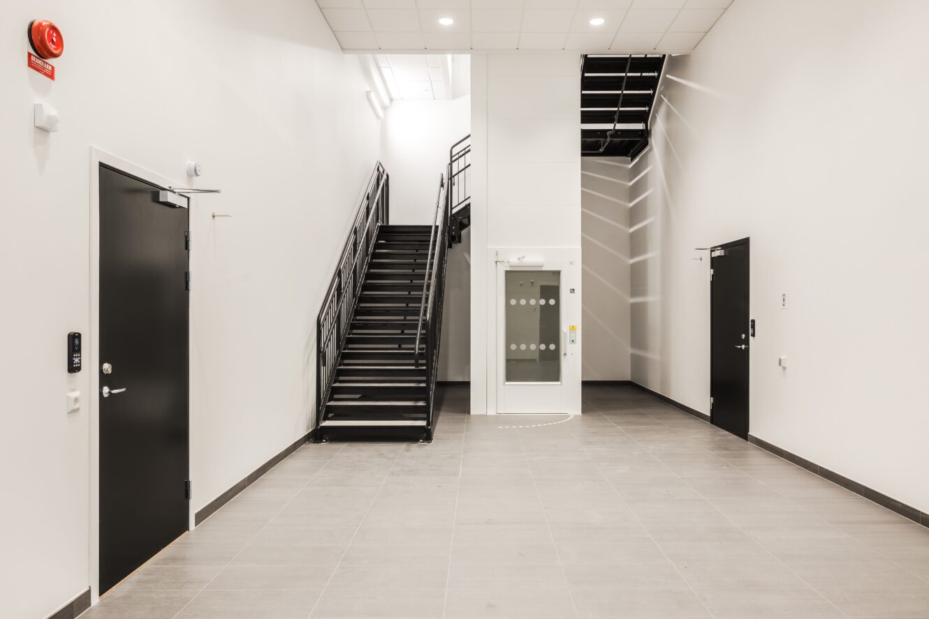 En ren, modern interiör med ljusgrått klinkergolv, vita väggar, svarta dörrar och en svart metalltrappa som leder till en övre våning.