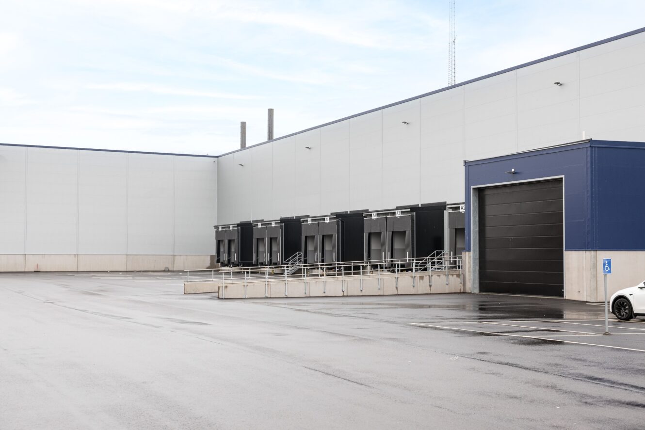 En modern lagerlokal med flera lastkajer, en stor garageport och en intilliggande parkeringsplats med markerade platser en molnig dag.