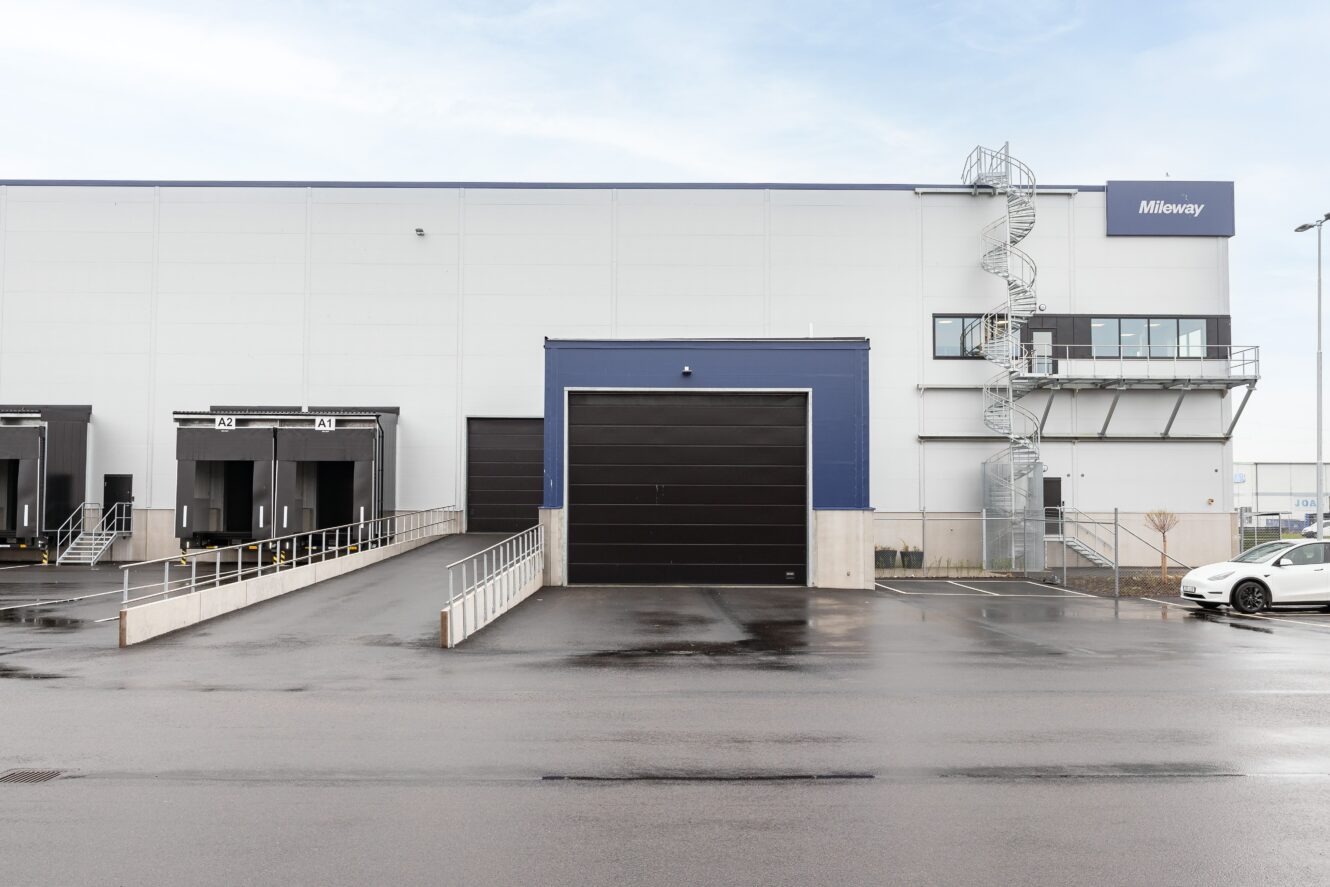 En modern lagerbyggnad med lastkaj, ramper, en spiraltrappa på sidan och en parkerad vit bil framför. Byggnaden har en skylt med texten "Midway".