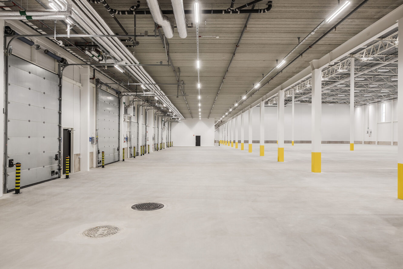 Vidvinkel över en tom lagerlokal med betonggolv, högt i tak, synliga rör och gula skyddsbarriärer på pelare. Stora garageportar finns på vänster sida.