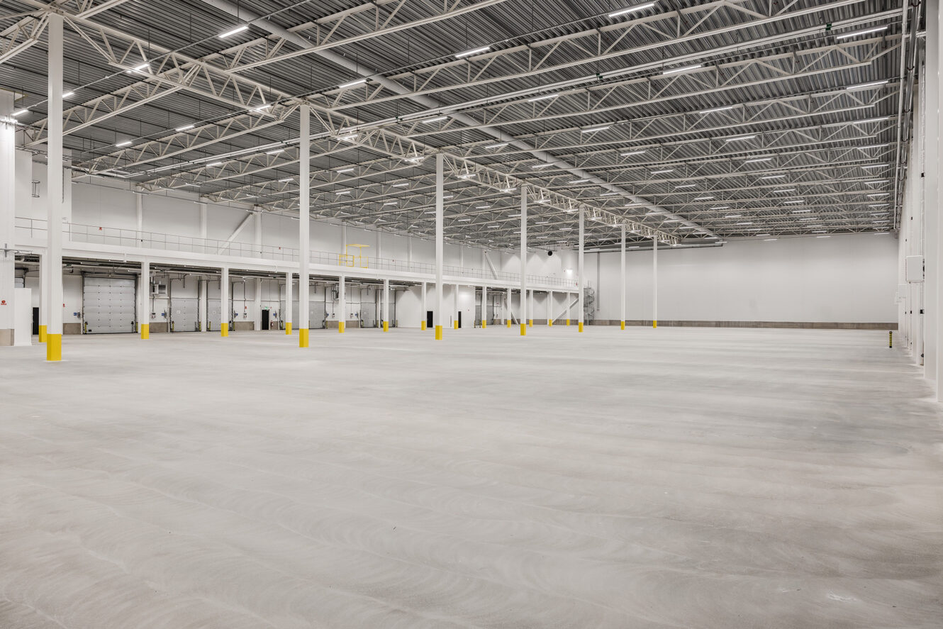 Stor tom lagerlokal med högt i tak, vita väggar, betonggolv, gula säkerhetspollare och industriell takbelysning.