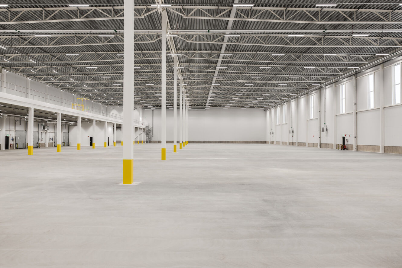 Rymlig, tom industrilokal med högt i tak, synliga bjälkar, stora fönster och gula säkerhetspollare på betonggolv.