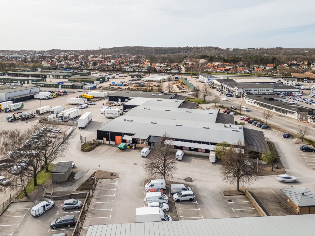 Flygfoto över ett industriområde med lagerlokaler, parkerade lastbilar, bilar och omgivande byggnader; bostadsområde syns i bakgrunden.