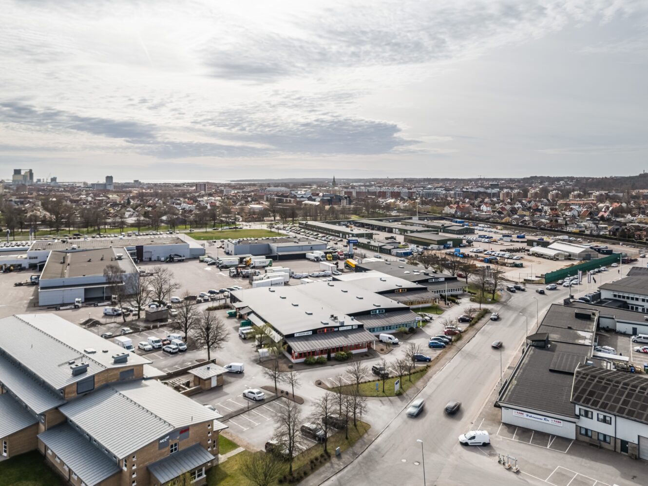 Flygfoto över ett affärs- och industriområde med flera låga byggnader, parkeringsplatser, parkerade bilar och en stadsbild i bakgrunden under en molnig himmel.