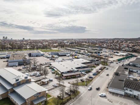 Flygfoto över ett affärs- och industriområde med flera låga byggnader, parkeringsplatser, parkerade bilar och en stadsbild i bakgrunden under en molnig himmel.