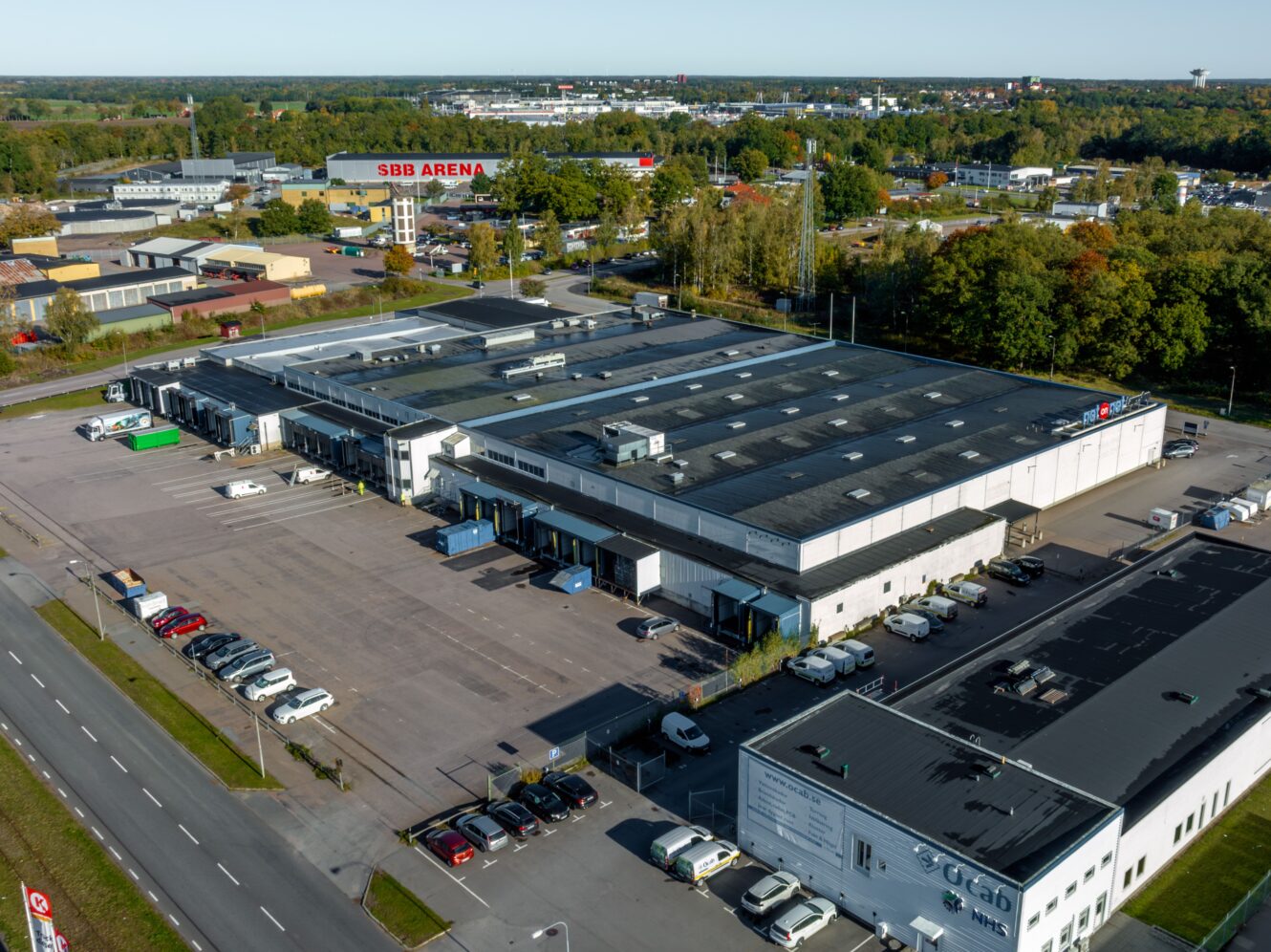 Flygfoto över ett industrikomplex med flera byggnader, parkeringsplatser och bilar; en skylt med texten SBB ARENA i bakgrunden bland träd och vägar.