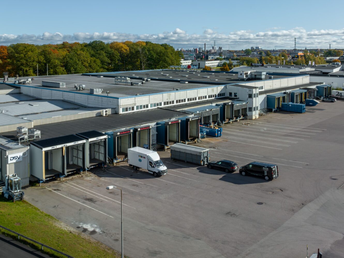 En stor lagerlokal med flera lastkajer, flera lastbilar och bilar parkerade utanför och träd i bakgrunden under en delvis molnig himmel.