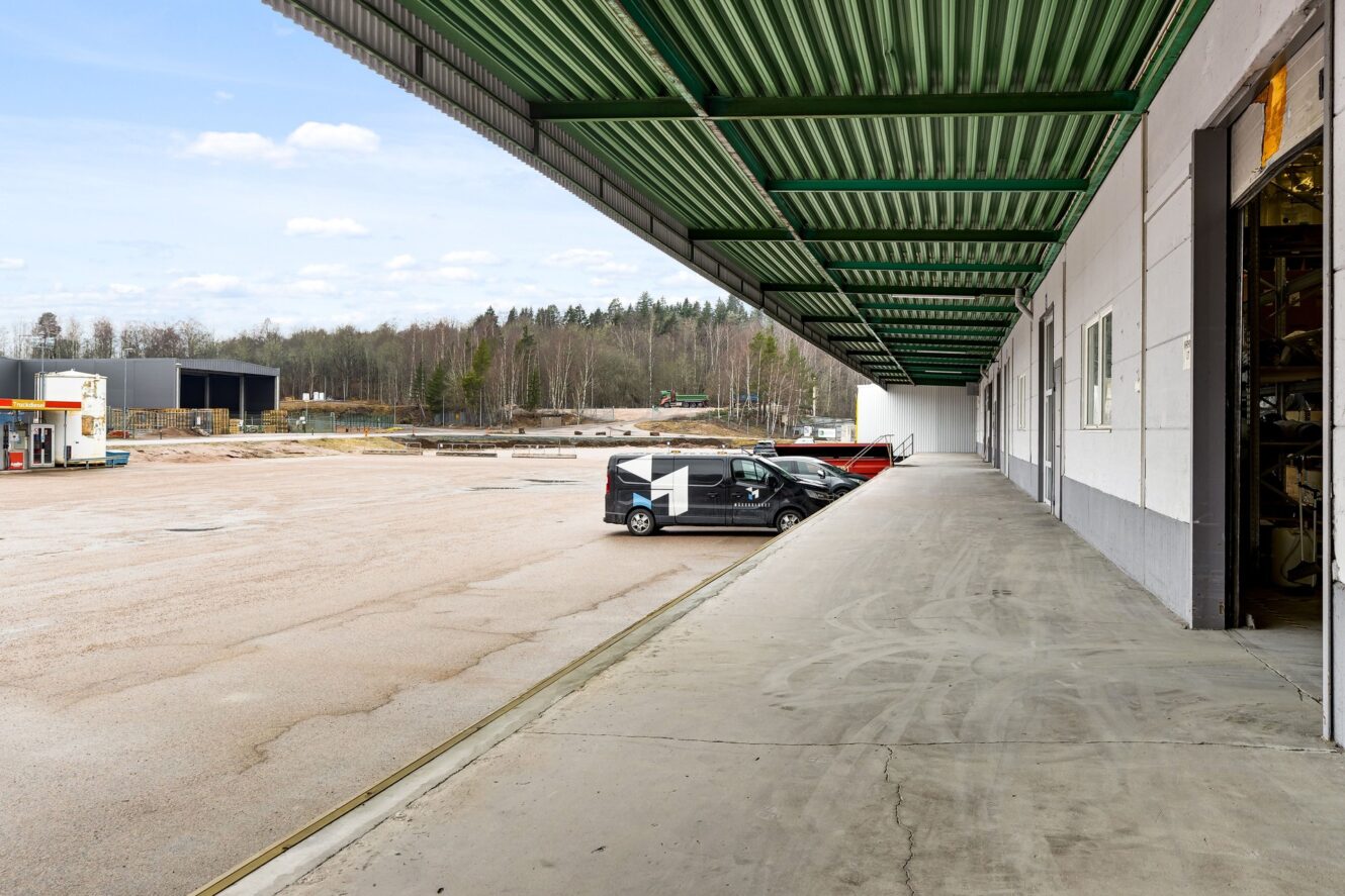 En lastkaj i betong med ett stort grönt skärmtak i metall, en parkerad skåpbil med siffran 1 på sidan och en industrigård med träd i bakgrunden.