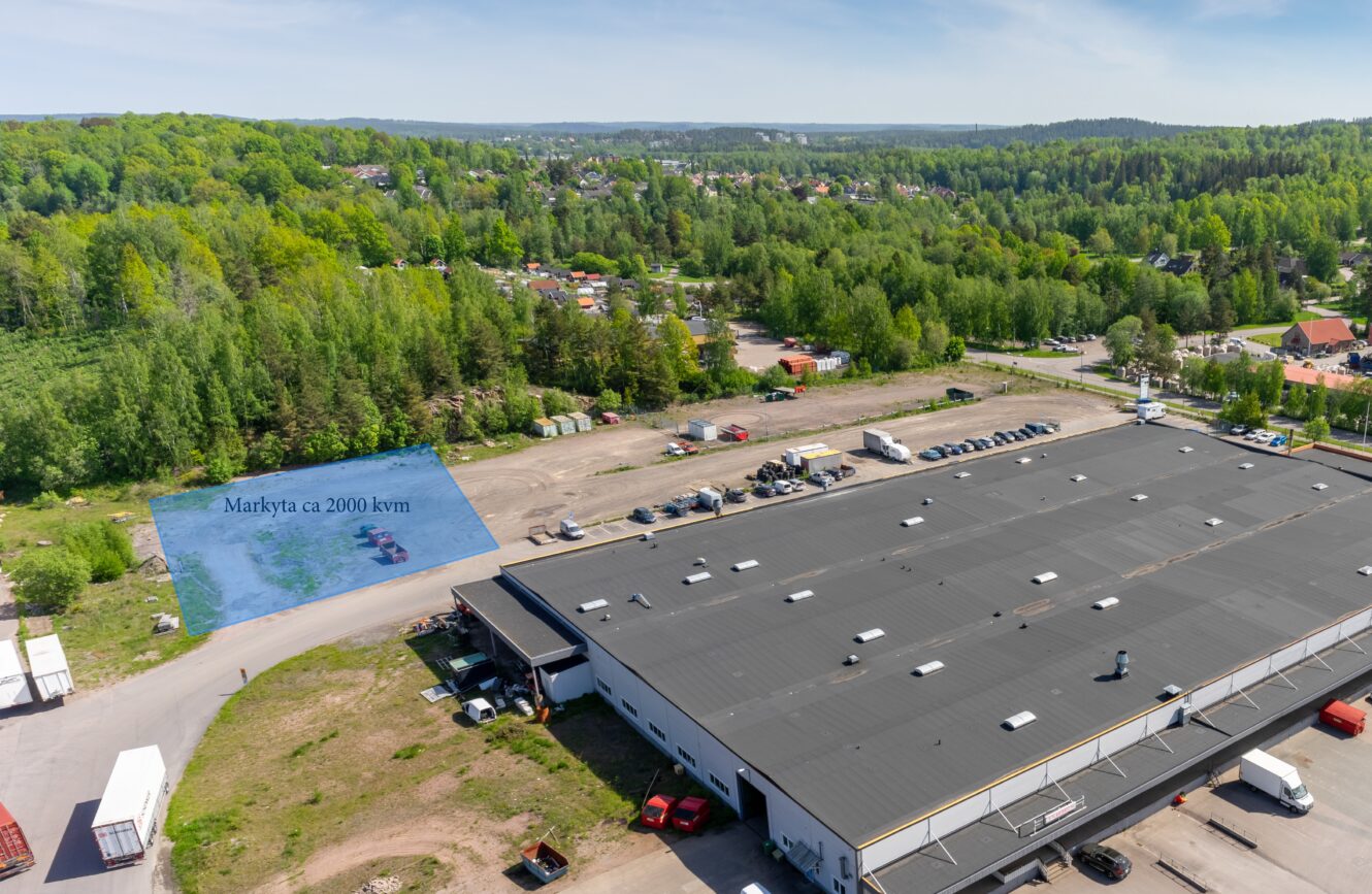 Flygfoto över ett industriområde med en stor lagerbyggnad i förgrunden och ett markerat utomhusområde på cirka 2000 kvadratmeter, omgivet av grönska och avlägsna hus.