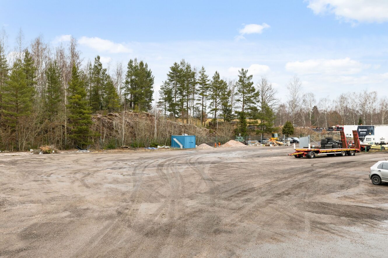 En grusplan med utspridda fordon, byggmaterial och en blå container, omgiven av träd under en delvis molnig himmel.