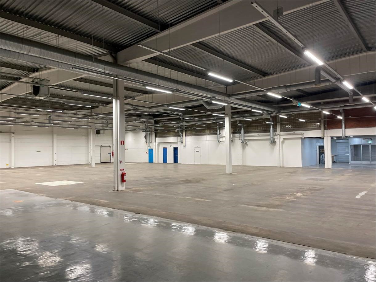 Rymlig, tom lagerlokal med polerade betonggolv, synliga rör i taket, stödpelare och väggar med dörrar och fönster. Stark takbelysning är synlig.