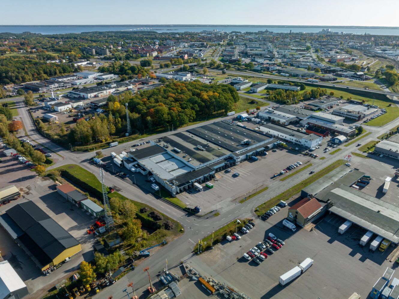 Flygbild över lagerlokaler, parkeringsplatser, vägar och träd i ett industriområde, med stadsbild och vatten i bakgrunden.