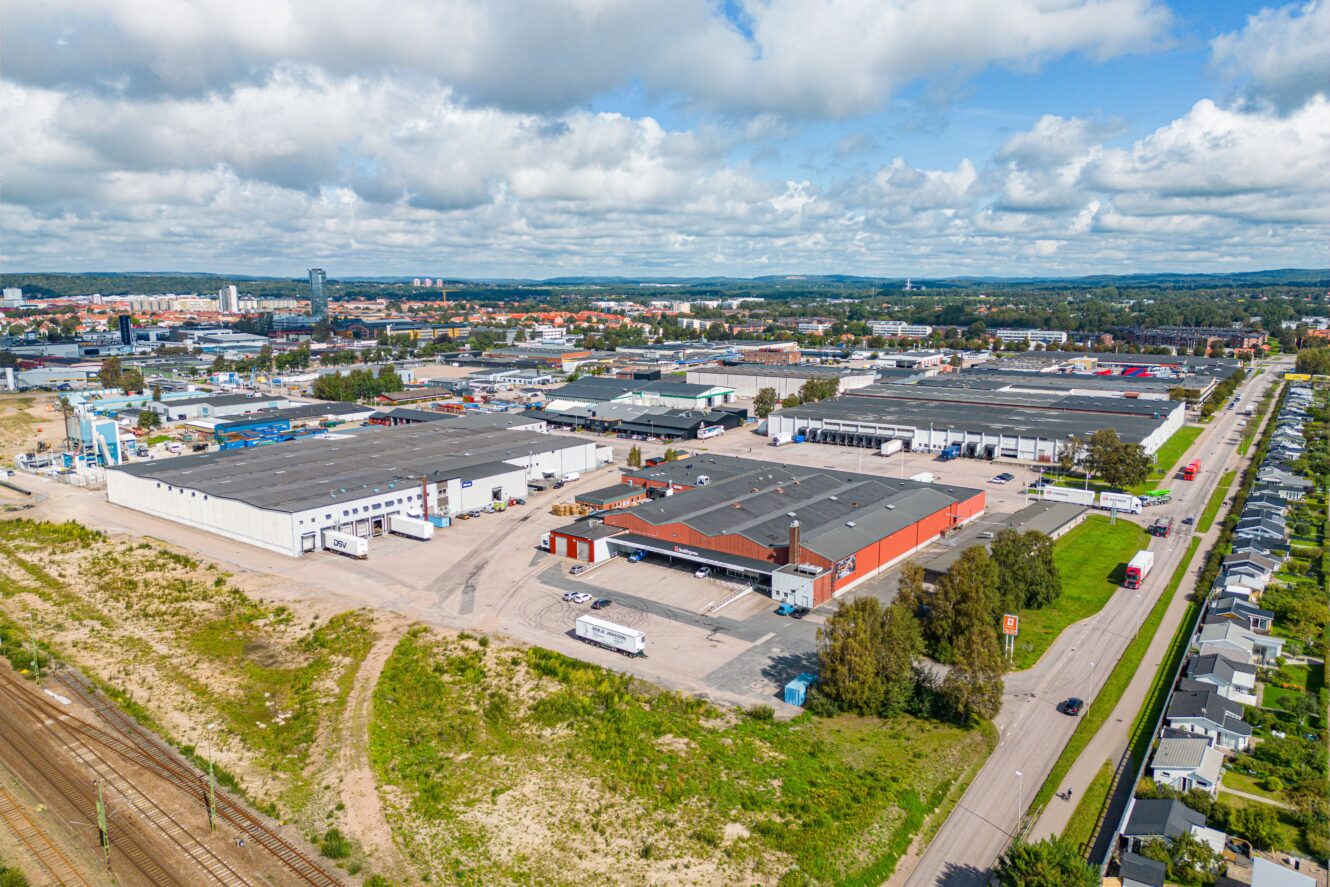 Flygfoto över ett industriområde med lagerbyggnader, lastbilar, parkeringsplatser, en närliggande järnväg och bostadshus längs en väg; stadsbild och grönska syns i bakgrunden.