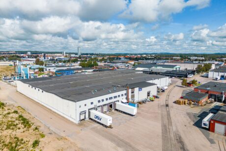 Flygfoto över ett industriområde med en stor lagerlokal, flera lastkajer, lastbilar parkerade utanför och närliggande byggnader under en delvis molnig himmel.