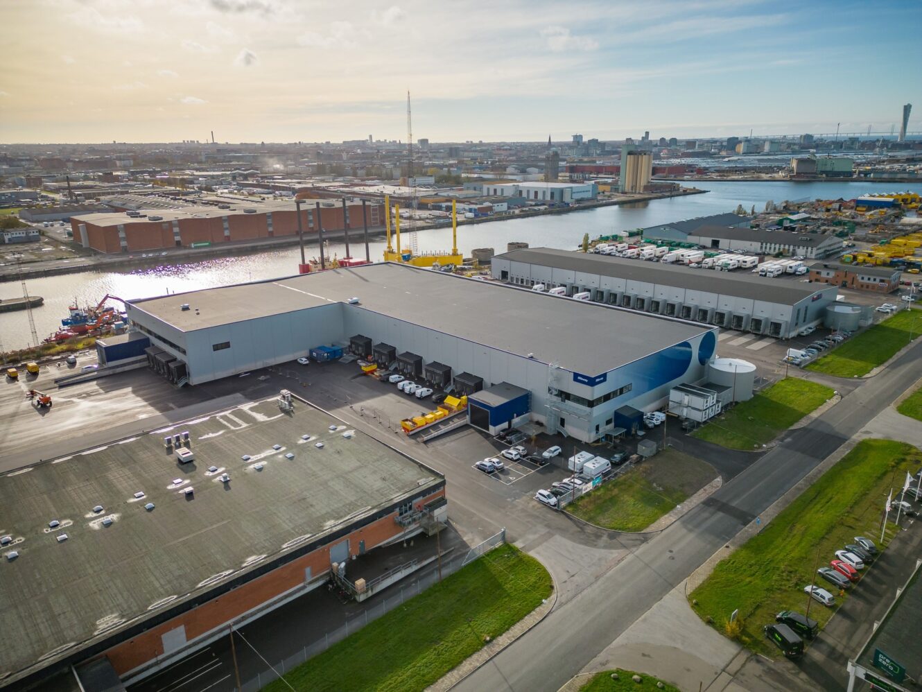 Flygfoto över ett industriellt lagerkomplex med intilliggande lastkajer, lastbilar och en närliggande vattenväg i ett urbant hamnområde under en delvis molnig himmel.