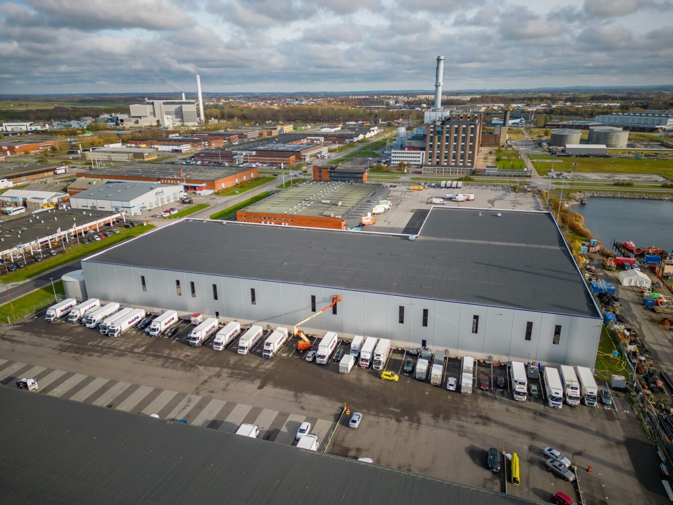En stor industrihall med flera lastbilar parkerade utanför, omgiven av andra industribyggnader och fabriker under en molnig himmel.