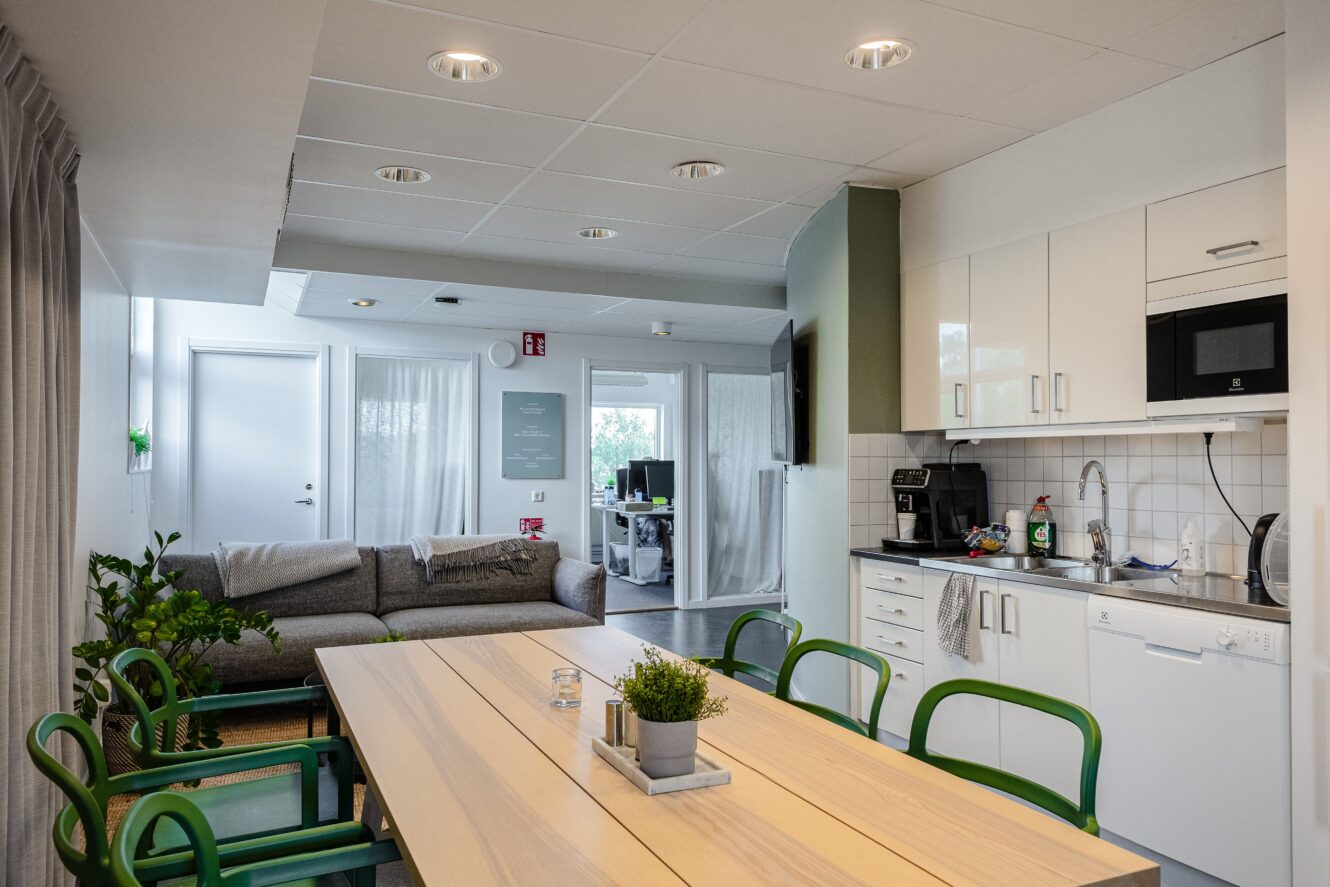 Modernt kök och vardagsrum med ett matbord i trä, gröna stolar, en grå soffa, vita skåp och vitvaror. Ett litet kontorsutrymme syns i bakgrunden.