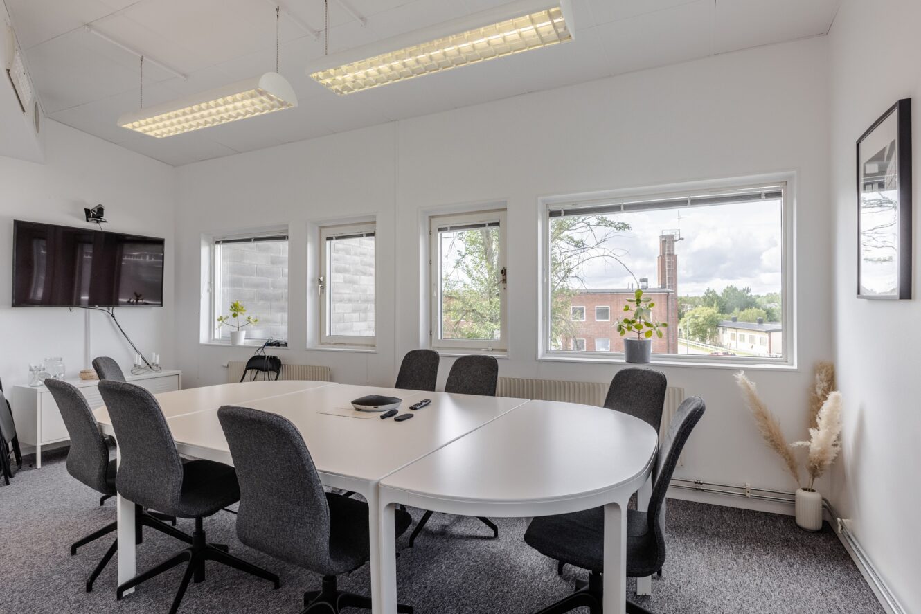 Ett modernt konferensrum med ett vitt bord, åtta gråa stolar, tre fönster, en väggmonterad TV och minimala dekorationer.