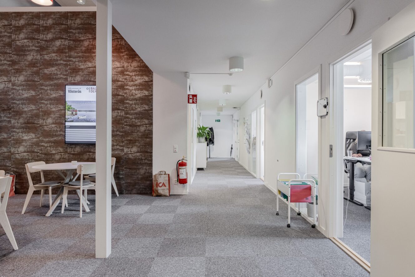 Modern kontorskorridor med heltäckningsmatta, vita väggar, flera dörrar, en liten matplats med bord och stolar, en brandsläckare och en shoppingväska på golvet.