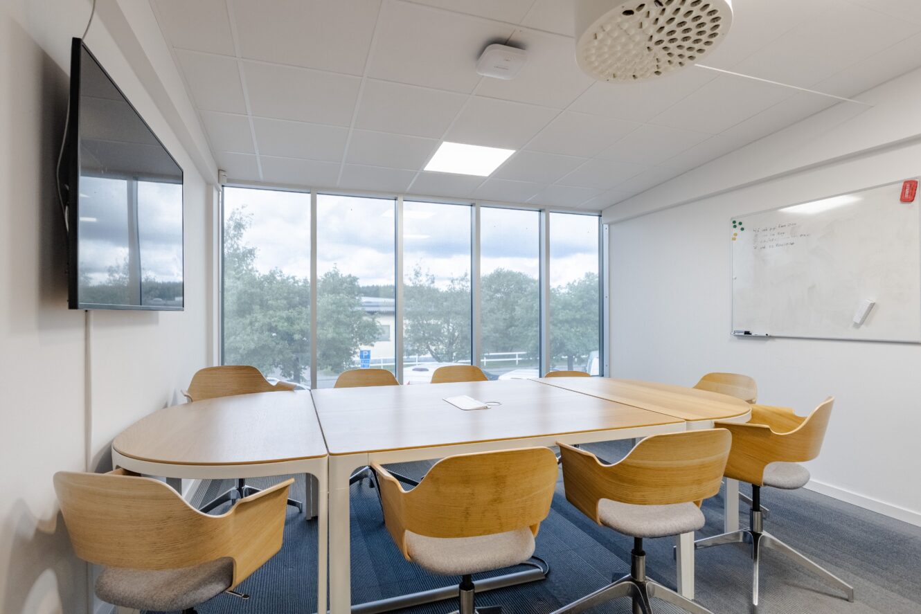 Ett modernt konferensrum med ett bord, åtta trästolar, en väggmonterad TV, stora fönster och en whiteboardtavla på väggen.