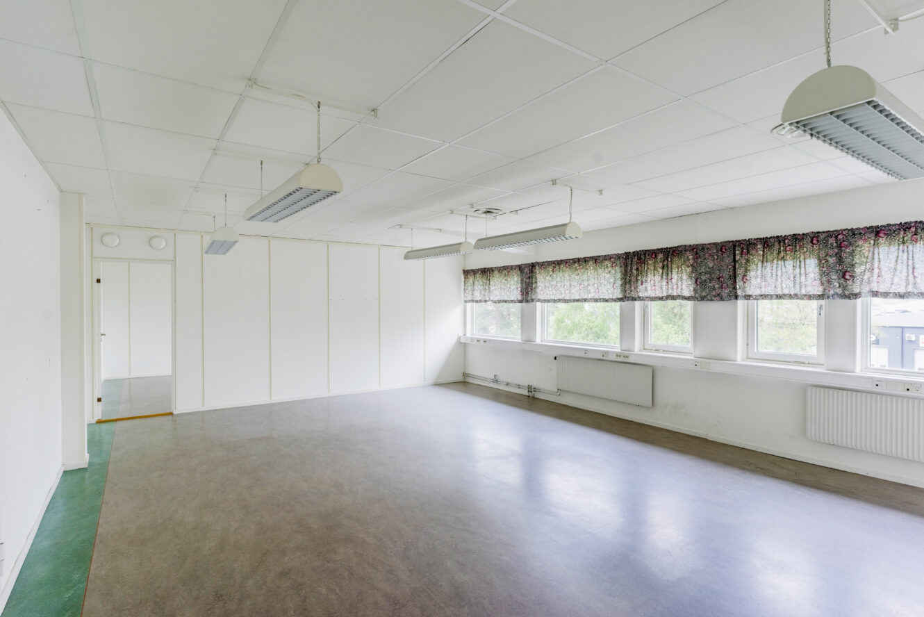 Tomt klassrum med vita väggar, stora fönster med blommiga gardiner, taklampor och ett grått linoleumgolv.