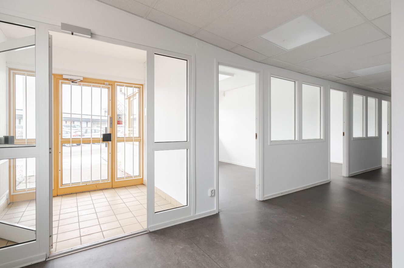 En korridor med flera små kontor med glasfönster och dörrar, som leder till en entré med en gallerförsedd ytterdörr och klinkergolv.