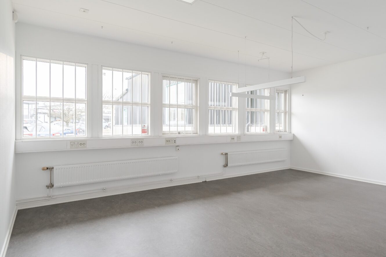 Tomt rum med vita väggar, stora fönster med spröjs, grått golv, element längs väggen och en hängande rektangulär armatur.