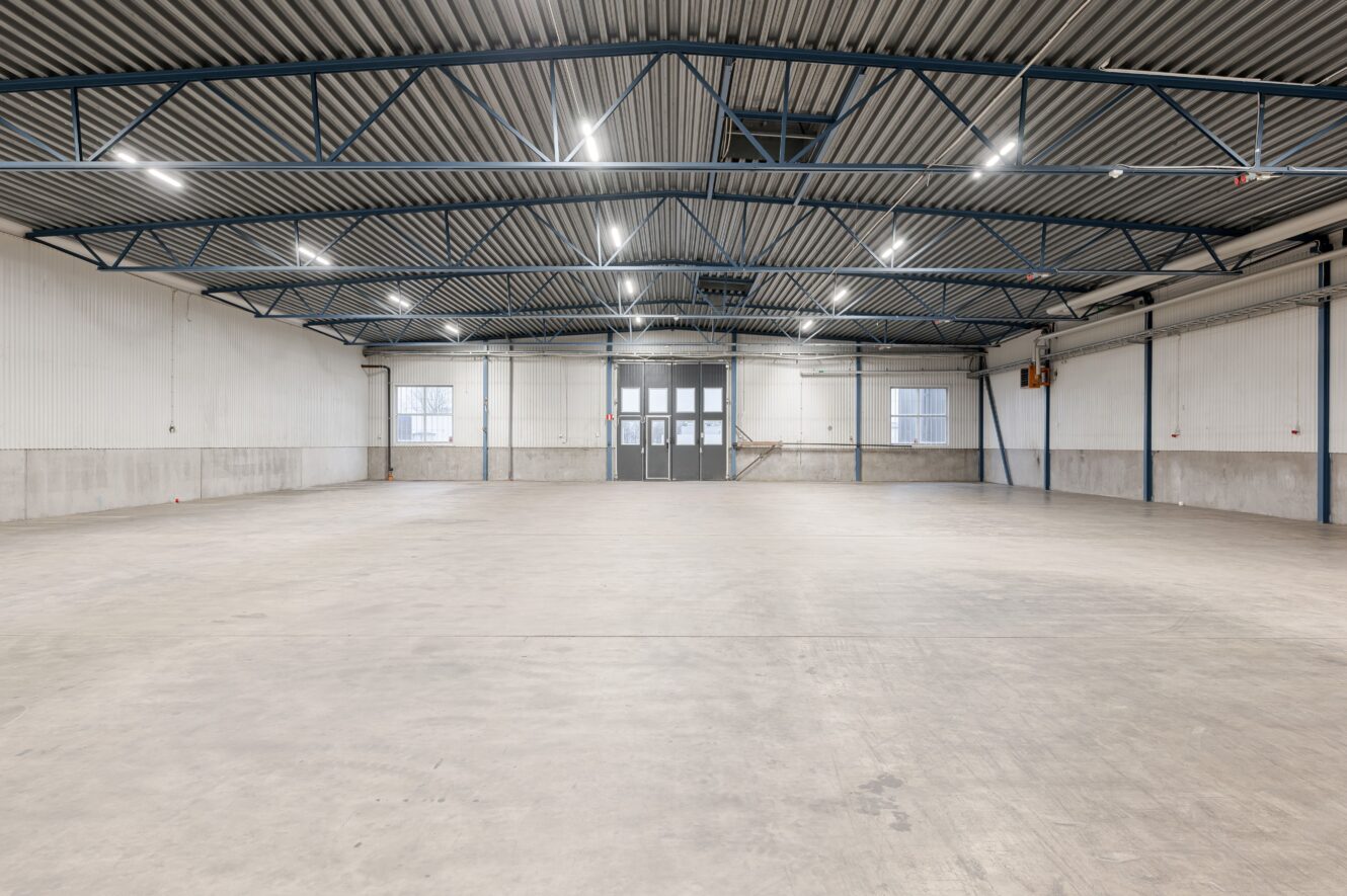 Rymlig, tom industrilokal med betonggolv, vita väggar, högt metalltak, synliga bjälkar och stora dubbeldörrar längst in.