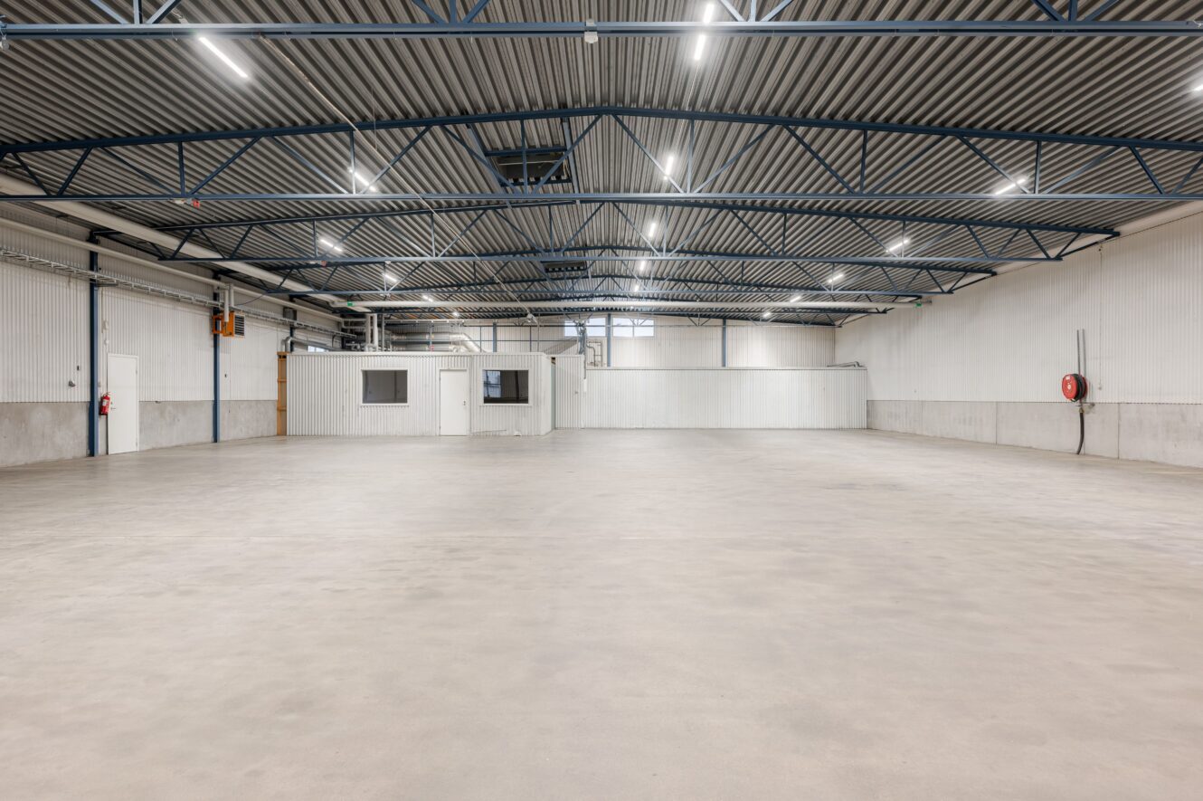 Tomt industrilager med betonggolv, högt metalltak, stark takbelysning och en liten vit kontorsbyggnad längs ena väggen.