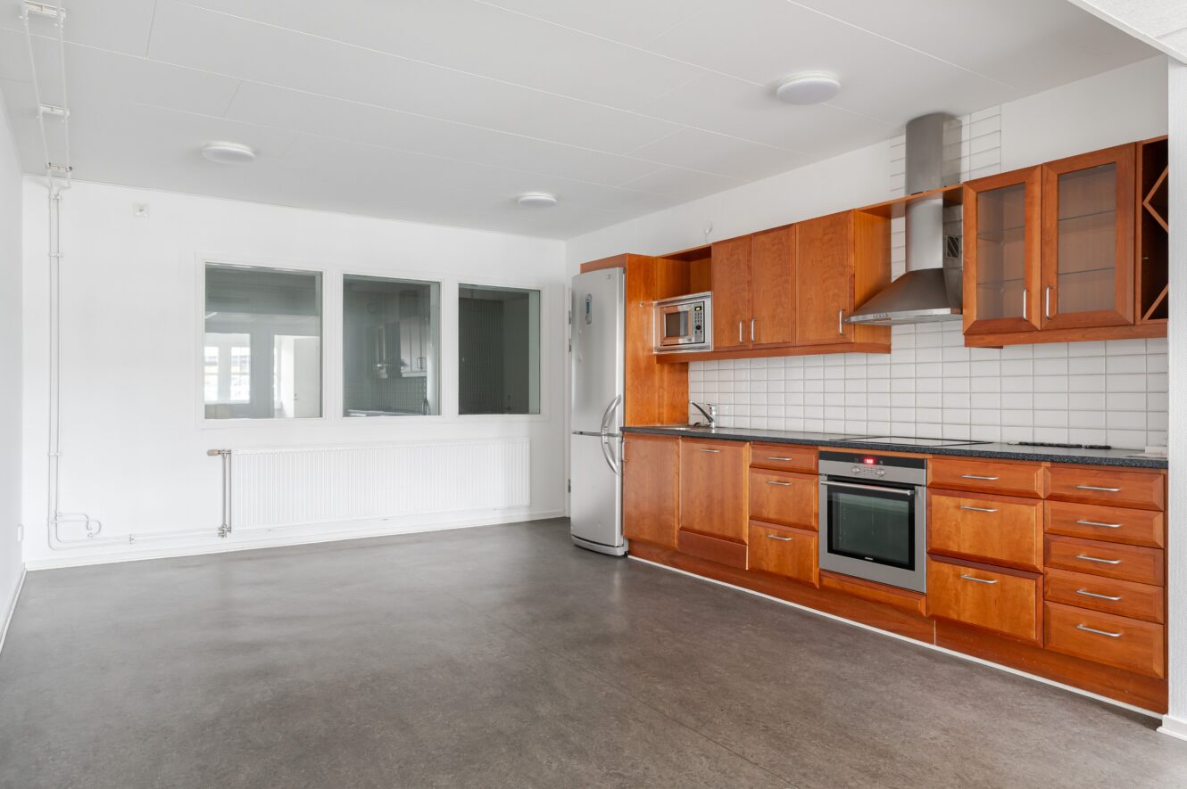 Modernt kök med skåp i trä, rostfria vitvaror, vitt kakel, grått golv och stora fönster i ett ljust, omöblerat rum.