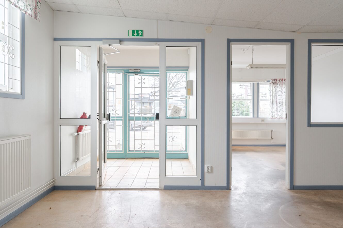 En ren, tom hall med klinkergolv, dubbeldörrar i glas som leder ut och en öppen dörr till ett ljust angränsande rum.