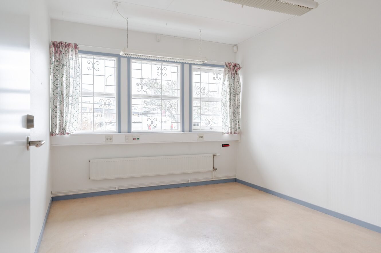 Tomt, omöblerat rum med vita väggar, stort fönster med mönstrade gardiner, element under fönstret och armaturer i taket.