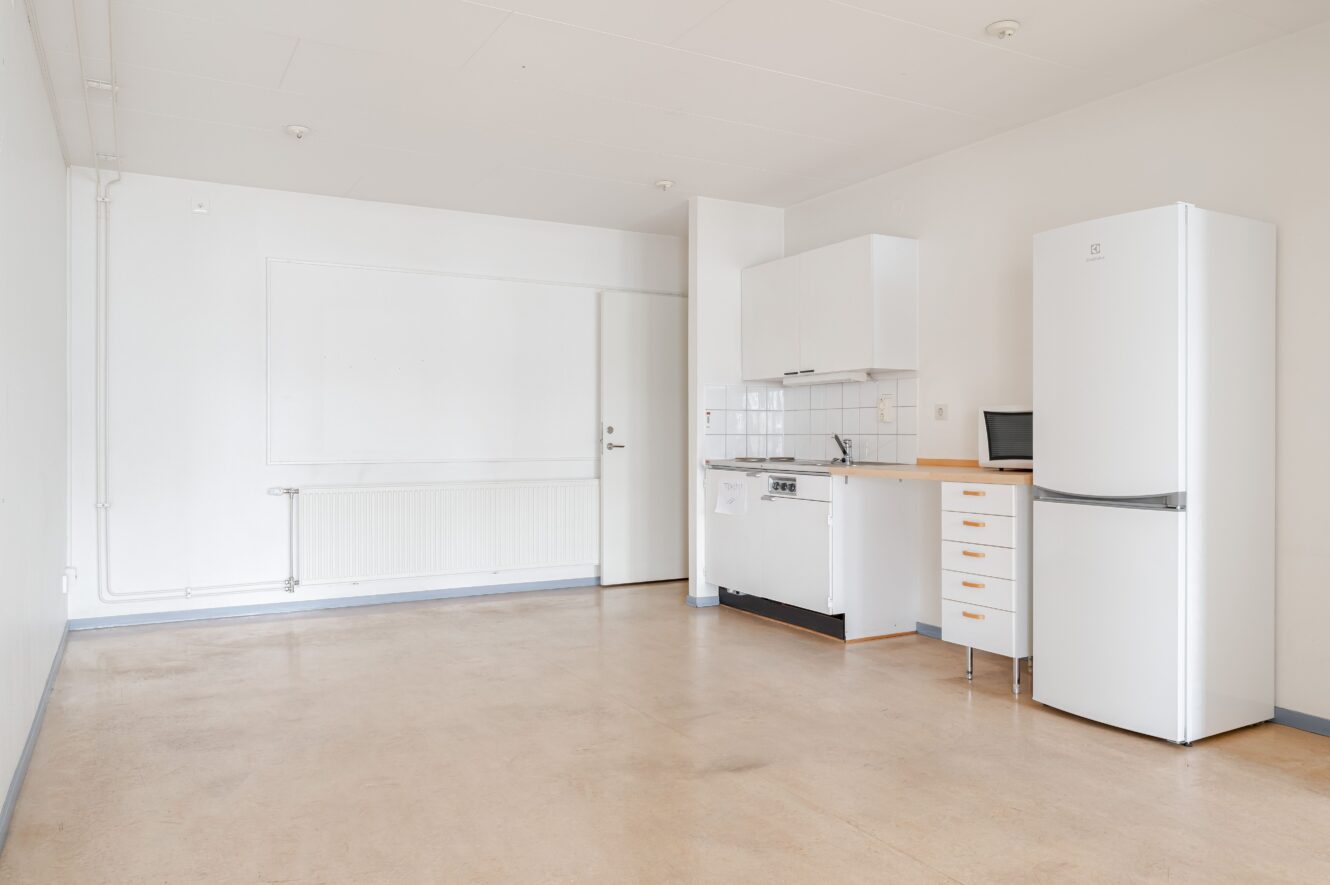 Minimalistiskt kök i ett vitt rum med kylskåp, mikrovågsugn, liten bänkskiva, diskho och skåp. Det finns gott om öppen yta och neutralt golv.