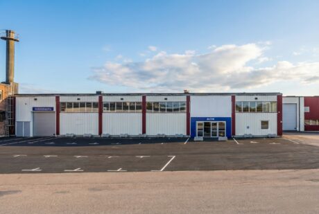 En stor industribyggnad med en blå butiksentré märkt BUTIK och en i stort sett tom parkeringsplats framför, under en delvis molnig himmel.
