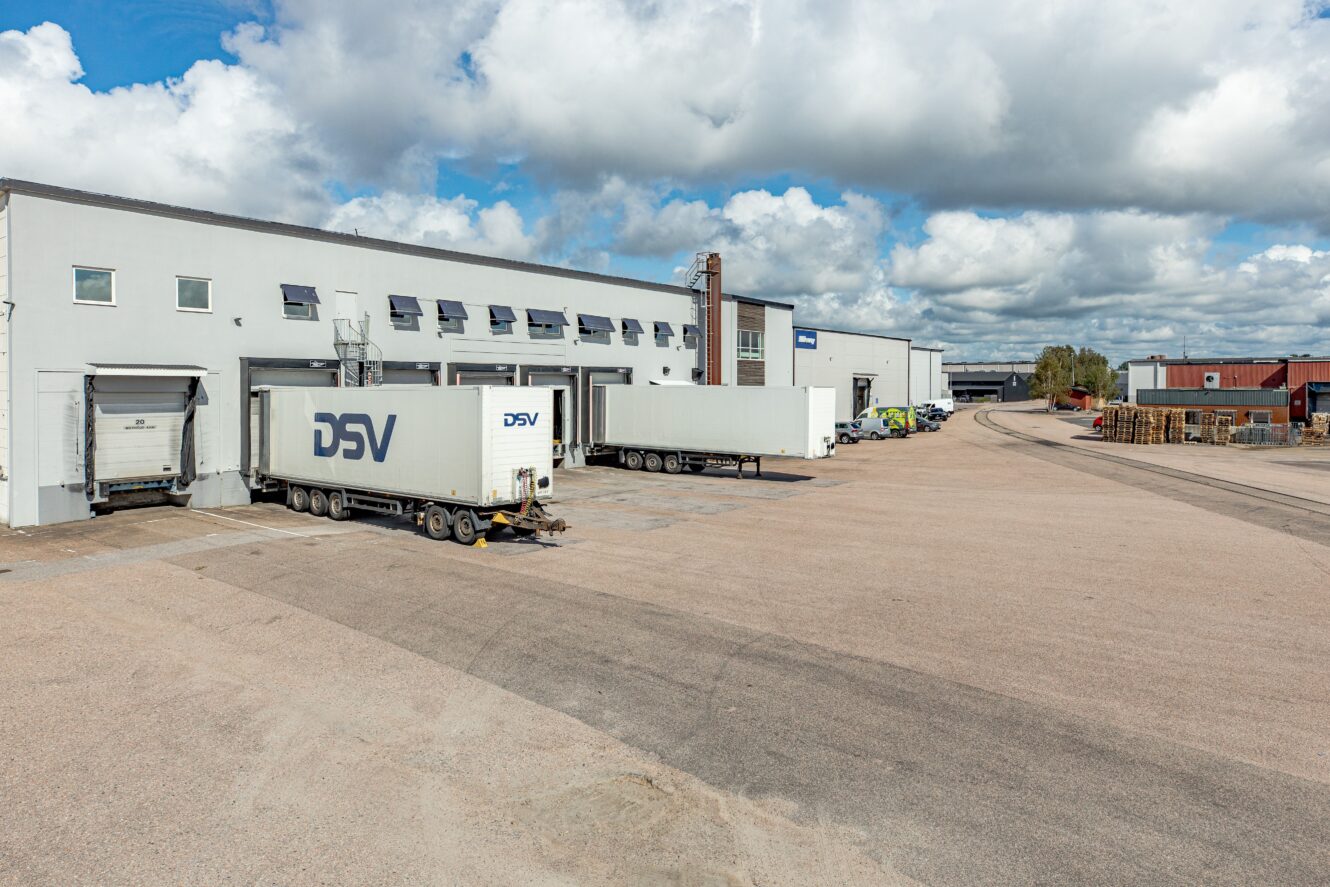 Flera vita lastsläp, varav ett är märkt DSV, står parkerade vid lastbryggor utanför ett industrilager under en delvis molnig himmel.