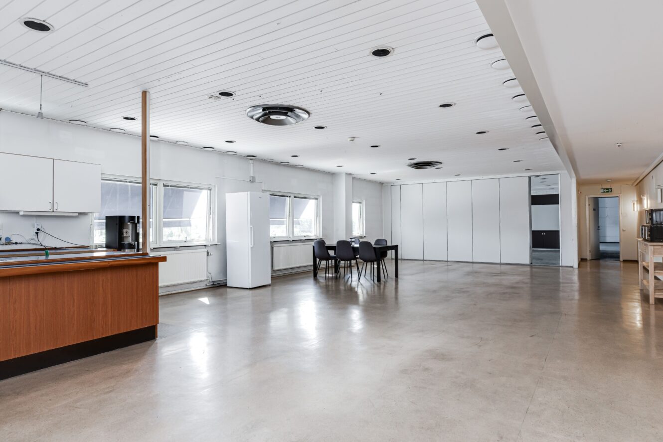 Rymligt, modernt fikarum med pentry, vita skåp, matbord med stolar, stora fönster och minimalistisk inredning.