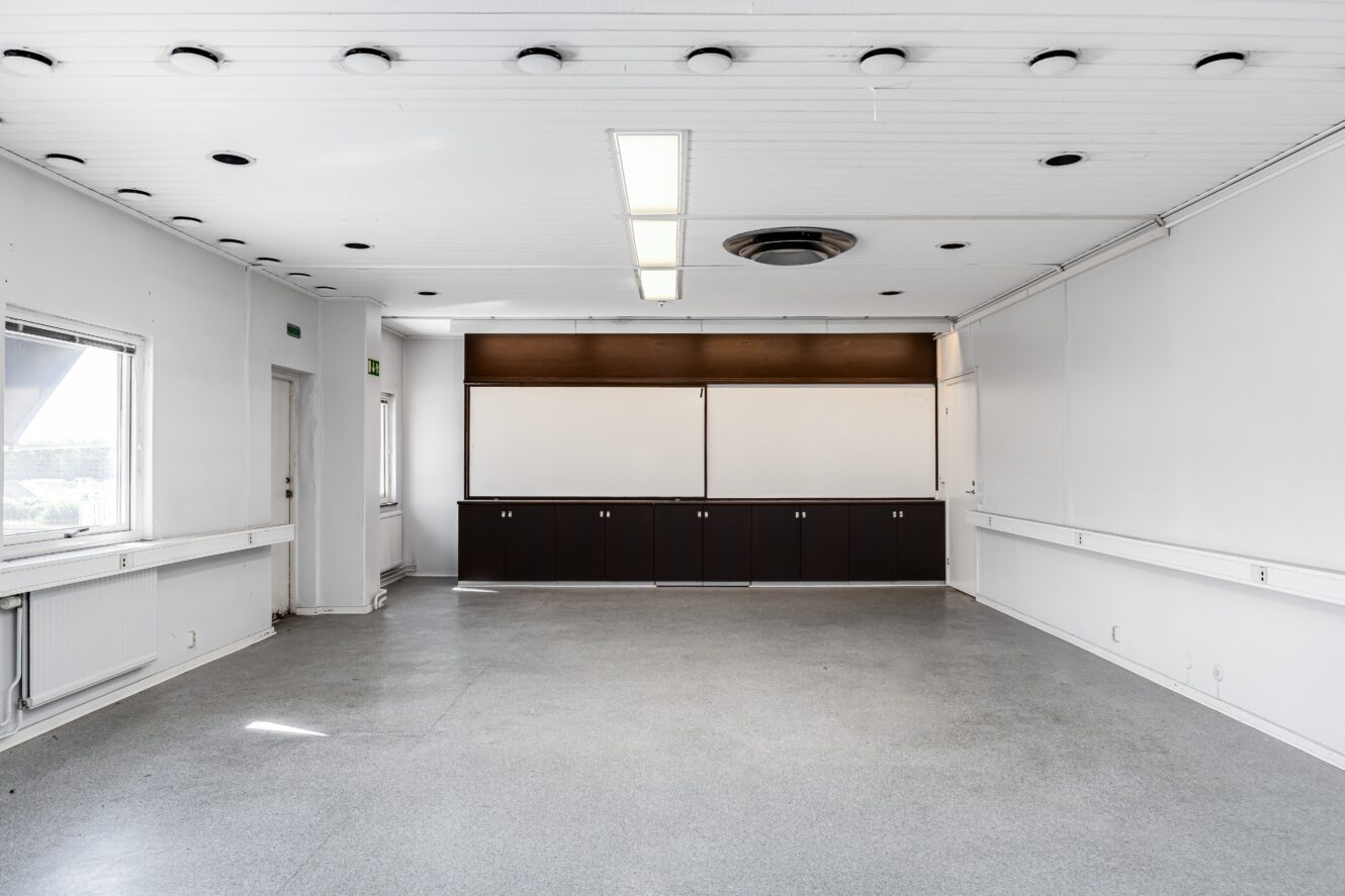 Ett tomt, väl upplyst rum med vita väggar, grått golv, ett stort fönster, takbelysning och ett inbyggt skåp med whiteboardtavlor längst bort.