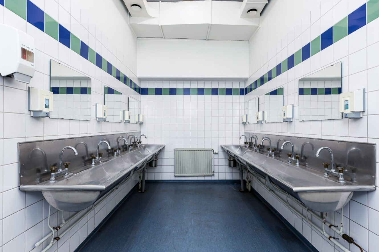 En offentlig toalett med två långa diskhoar i rostfritt stål, flera kranar, väggmonterade tvålbehållare, speglar och vita kaklade väggar med blå och gröna accenter.