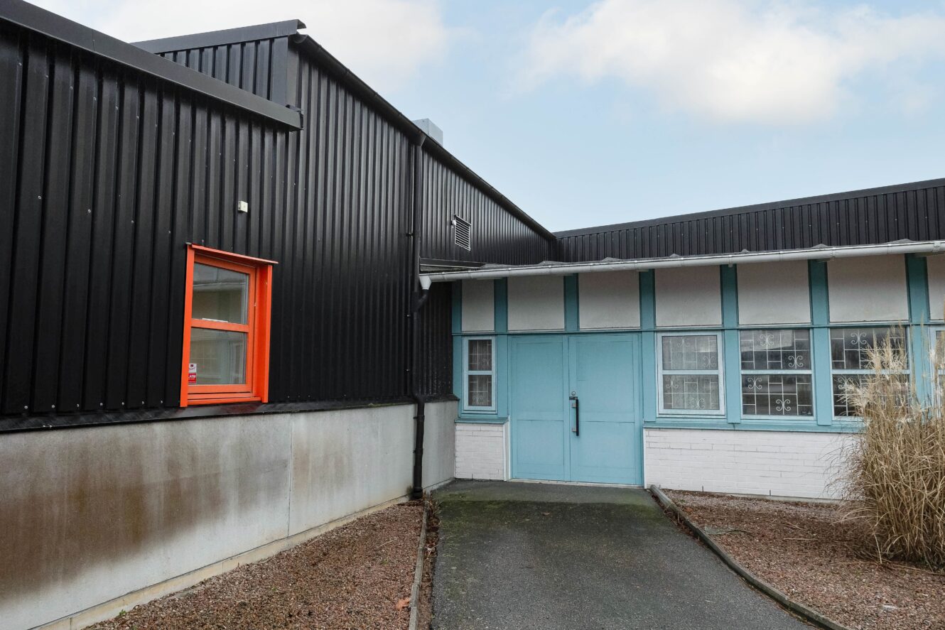 En modern byggnad med svart metallbeklädnad, ett ljusorange fönster, ljusblå dubbeldörrar och en grusplan.