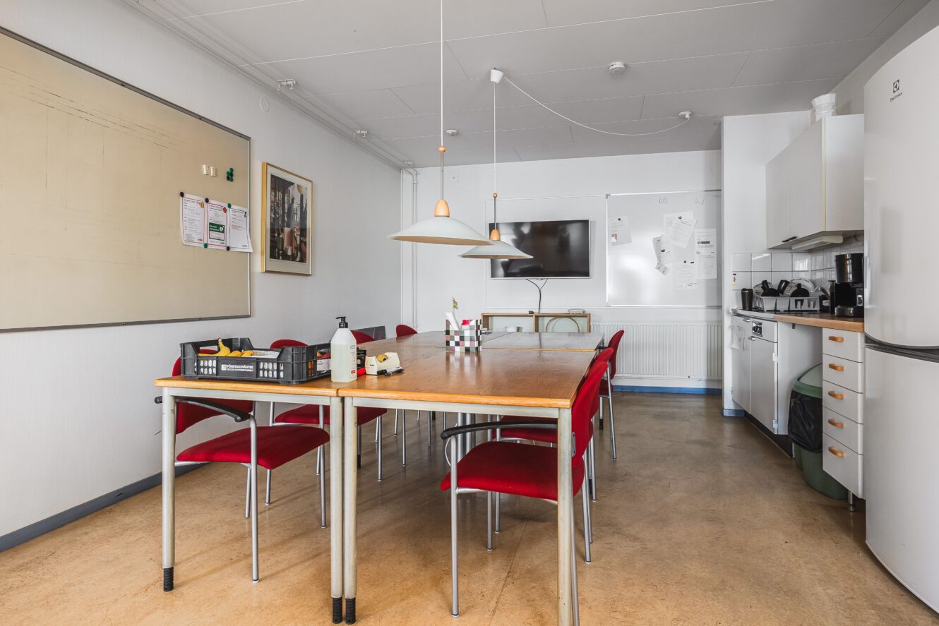 Ett fikarum för personalen med ett långt bord, röda stolar, whiteboard, väggmonterad TV, köksmaskiner och olika föremål på bord och bänkar.