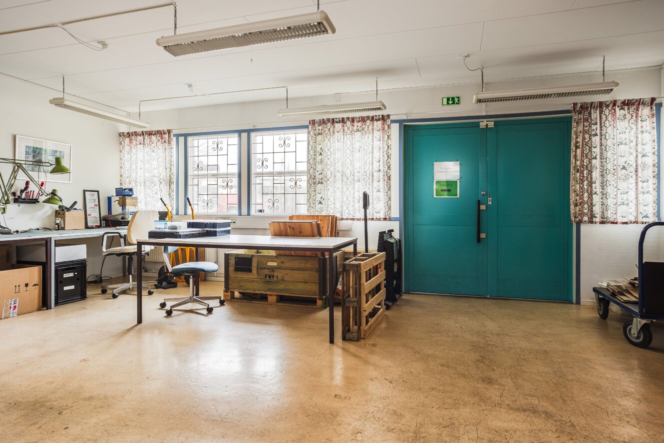 Rymlig, väl upplyst verkstad med två skrivbord, industribord, kontorsstolar och stora tealfärgade dubbeldörrar. Fönster med mönstrade gardiner släpper in naturligt ljus.
