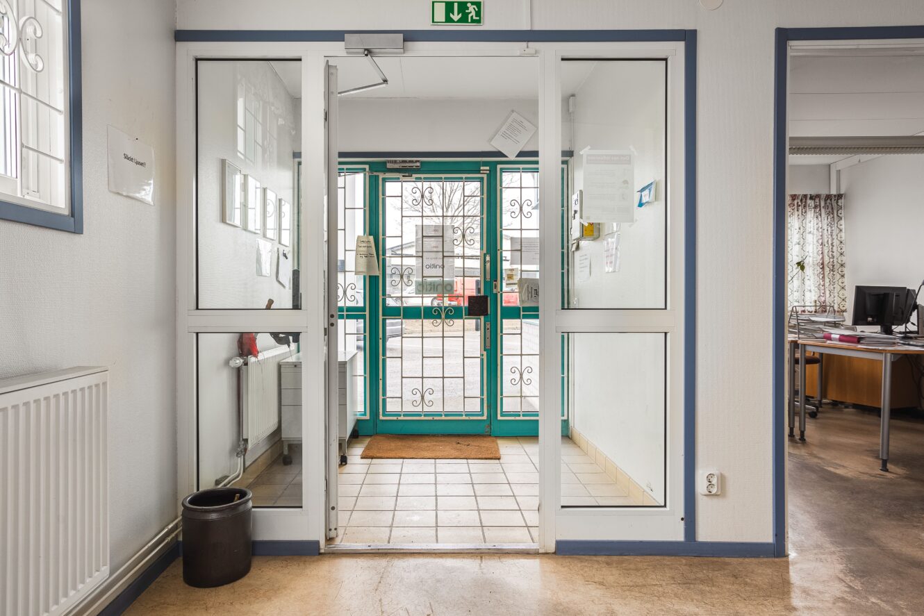 En dubbeldörr i glas med metallstänger leder till ett utomhusområde; interiören har vita väggar, klinkergolv och ett kontor som syns till höger.