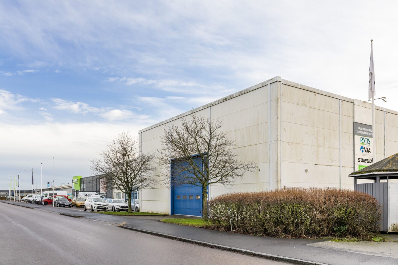 En vit industribyggnad med blå garageport och skyltning, belägen på en gata med parkerade bilar och lövlösa träd.