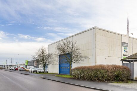 En vit industribyggnad med blå garageport och skyltning, belägen på en gata med parkerade bilar och lövlösa träd.