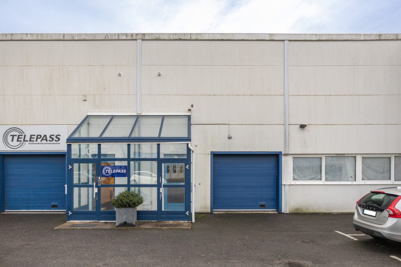 Huvudentrén till en beige industribyggnad med blå dörrar, en glasvestibul och en Telepass-skylt; en parkerad bil syns delvis till höger.