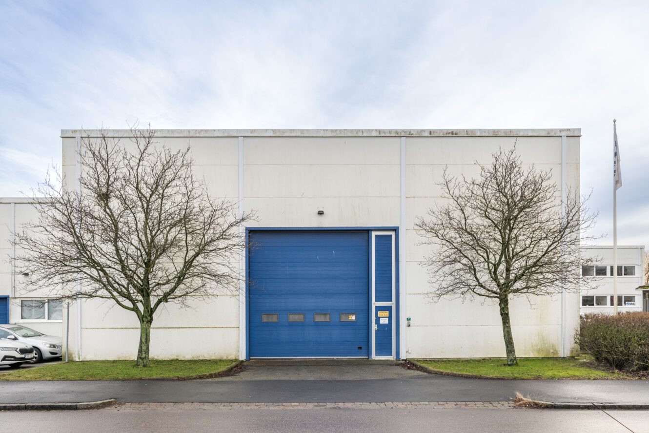 En vit industribyggnad med en stor blå garageport, två lövlösa träd på vardera sidan och parkerade bilar som delvis syns till vänster.