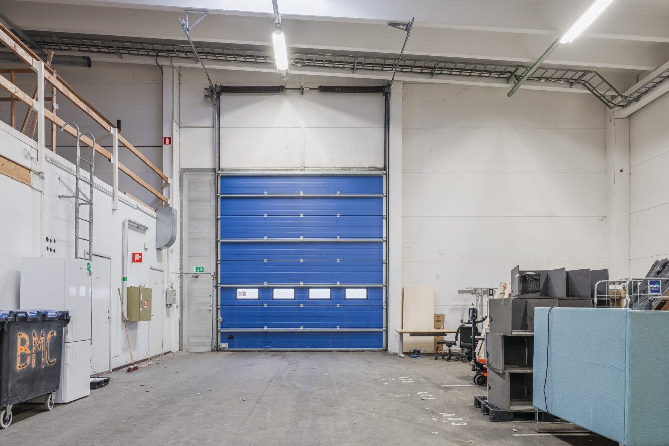 Inredning i ett industrilager med en stängd blå takskjutport, utspridd utrustning, soptunnor och olika föremål längs den högra väggen.