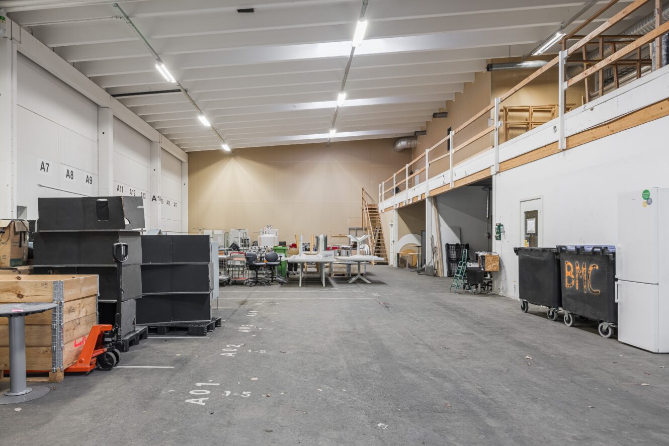 En rymlig industrilagerinredning med förvaringsfack, utrustning, arbetsbord och ett entresolplan med ett räcke längs ena väggen.