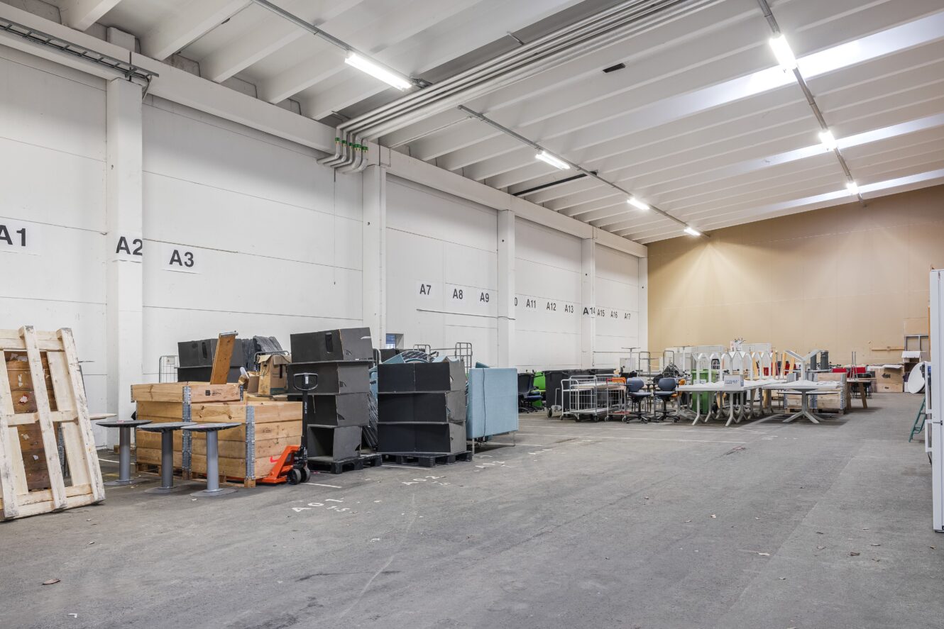 Stort industrilager med högt i tak, vita väggar med avdelningsnummer och diverse utrustning, pallar och möbler utspridda längs ena sidan.